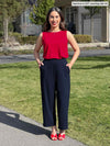 Miik model Yasmine (5'0", xsmall, petite) smiling with both hands on pocket wearing Miik's Ada reversible draped cowl neck tank in poppy red with a navy pant