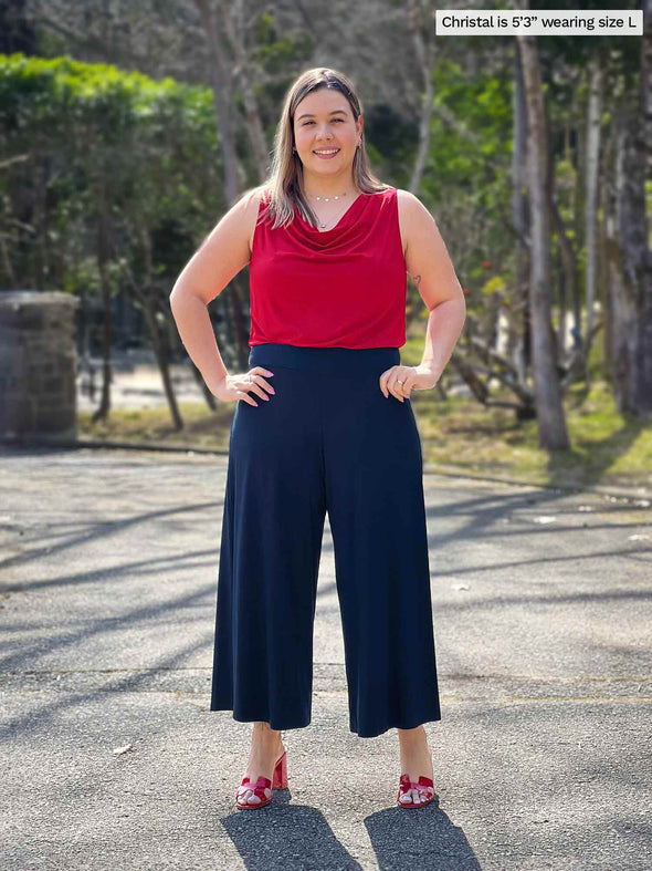 Miik model Christal (5'3", large) smiling wearing a flare navy pant along with Miik's Ada reversible draped cowl neck tank in poppy red