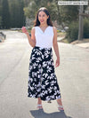 Miik model Yasmine (5'0, xsmall, petite) smiling wearing a printed flare pant along with Miik's Ada reversible draped cowl neck tank in white and high heels 