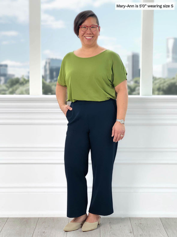 Miik model Mary-Ann (5'0", small) smiling while standing in front of a window wearing Miik's Adisa reversible dolman top in green moss with a navy pant 