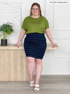 Miik model Bri (5'5", xlarge) leaning against to a coffee table wearing a striped navy skirt with Miik's Adisa reversible dolman top in green moss