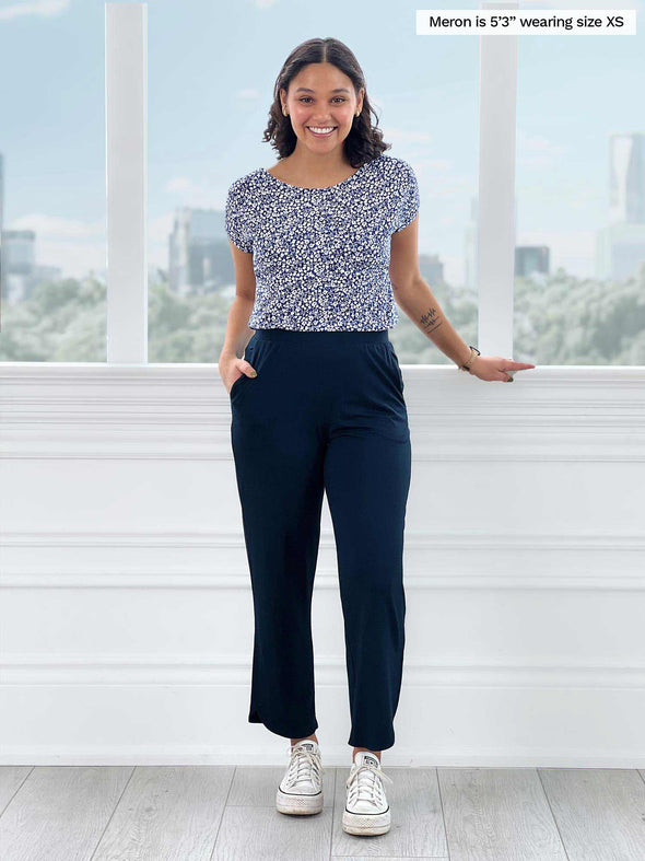 Miik model Meron (5'3", xsmall) smiling while standing in front of a window wearing Miik's Akira tulip hem capri pant in navy with a basic tee in baby's breath print and sneakers 