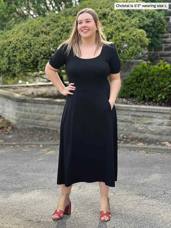 Miik model Christal (5'3", large) smiling with one hand on pocket wearing Miik's Anika puff sleeve midi dress in black 