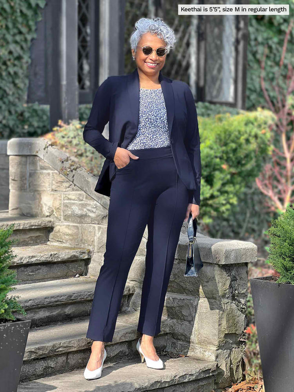 Miik model Keethai (5'5", medium) smiling wearing Miik's Christal pull-on pintuck ankle pant in regular length in navy with a blazer in the same colour and a printed tank