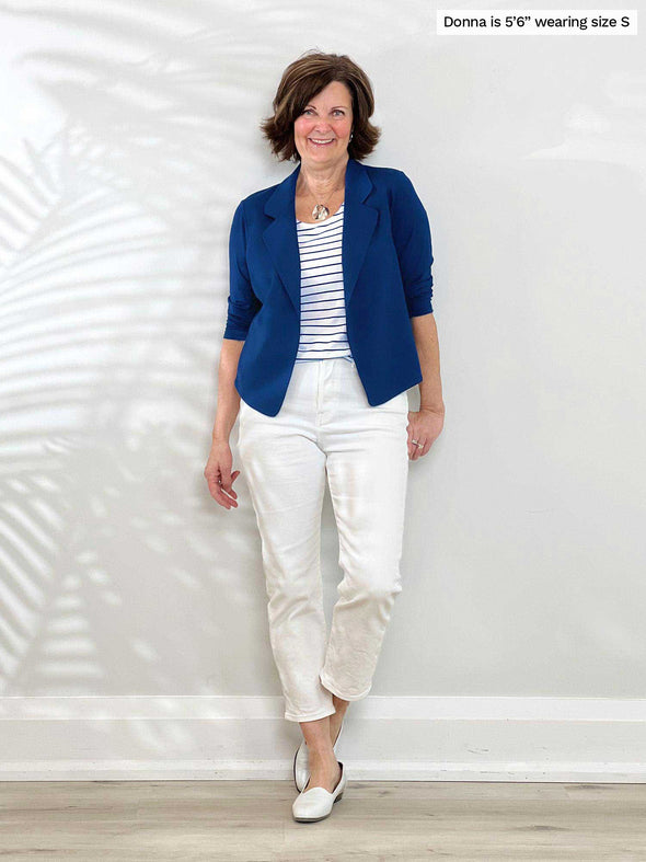 Miik founder Donna (5'6", small) smiling and leaning against to a white wall wearing Miik's Dorit boxy soft blazer in ink blue, a striped tank and white jeans 