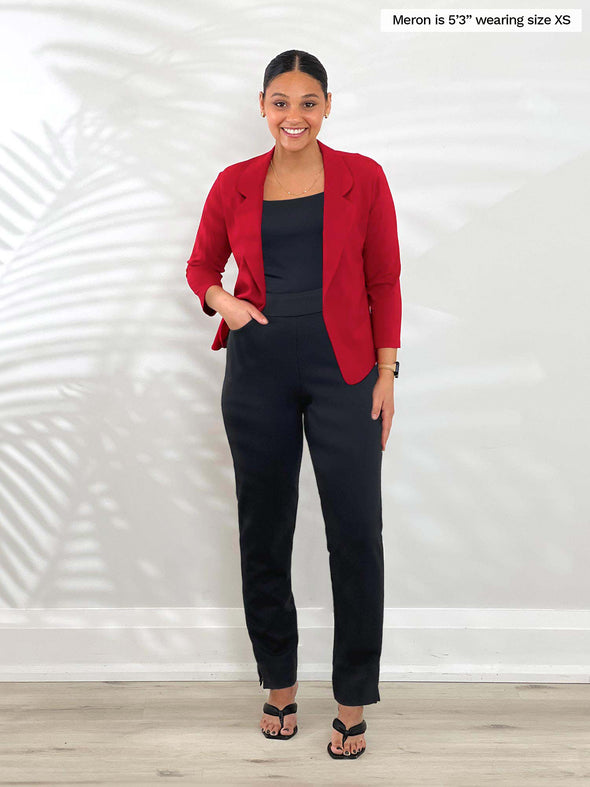 Miik model Meron (5'3", xsmall) smiling while standing in front of a white wall wearing Miik's Dorit boxy soft blazer in poppy red along with a black tank and pant 