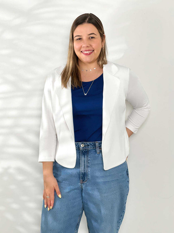 Miik model Christal (5'3", medium) smiling while standing in front of a white wall wearing Miik's Dorit boxy soft blazer in white with a ink blue tee and jeans 