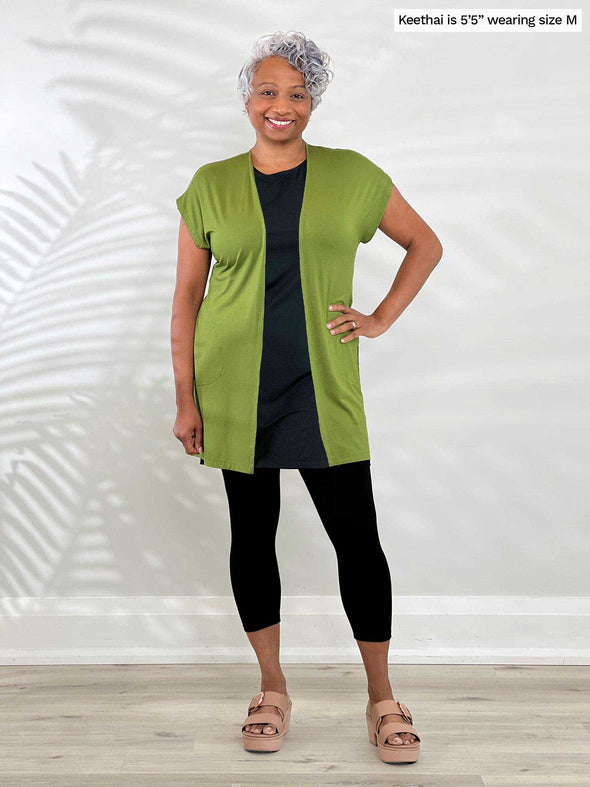 Miik model Keethai (5'5", medium) smiling while standing in front of a white wall wearing Miik's Evie short sleeve pocket cardigan in green moss with a capri legging and a black tunic