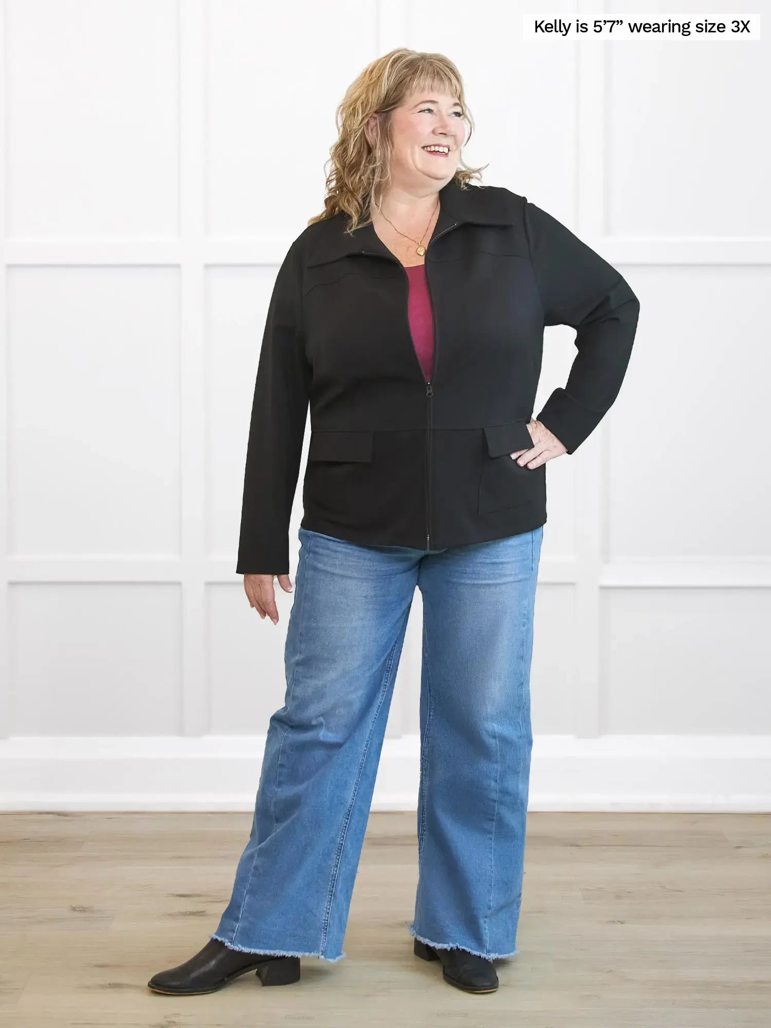 Woman wearing a black jacket and blue jeans standing against a white wall.