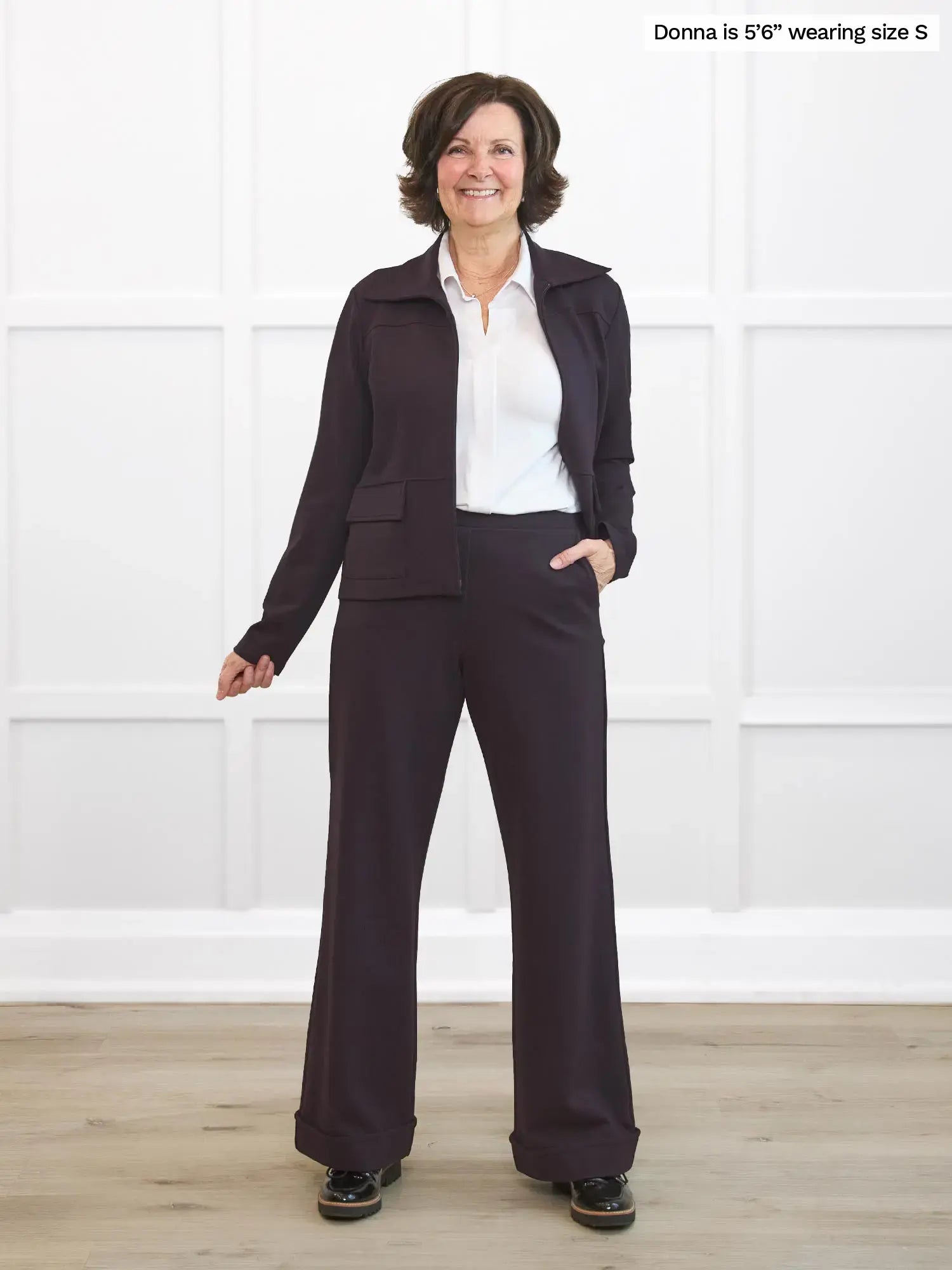 Woman wearing a dark brown suit standing against a white wall.