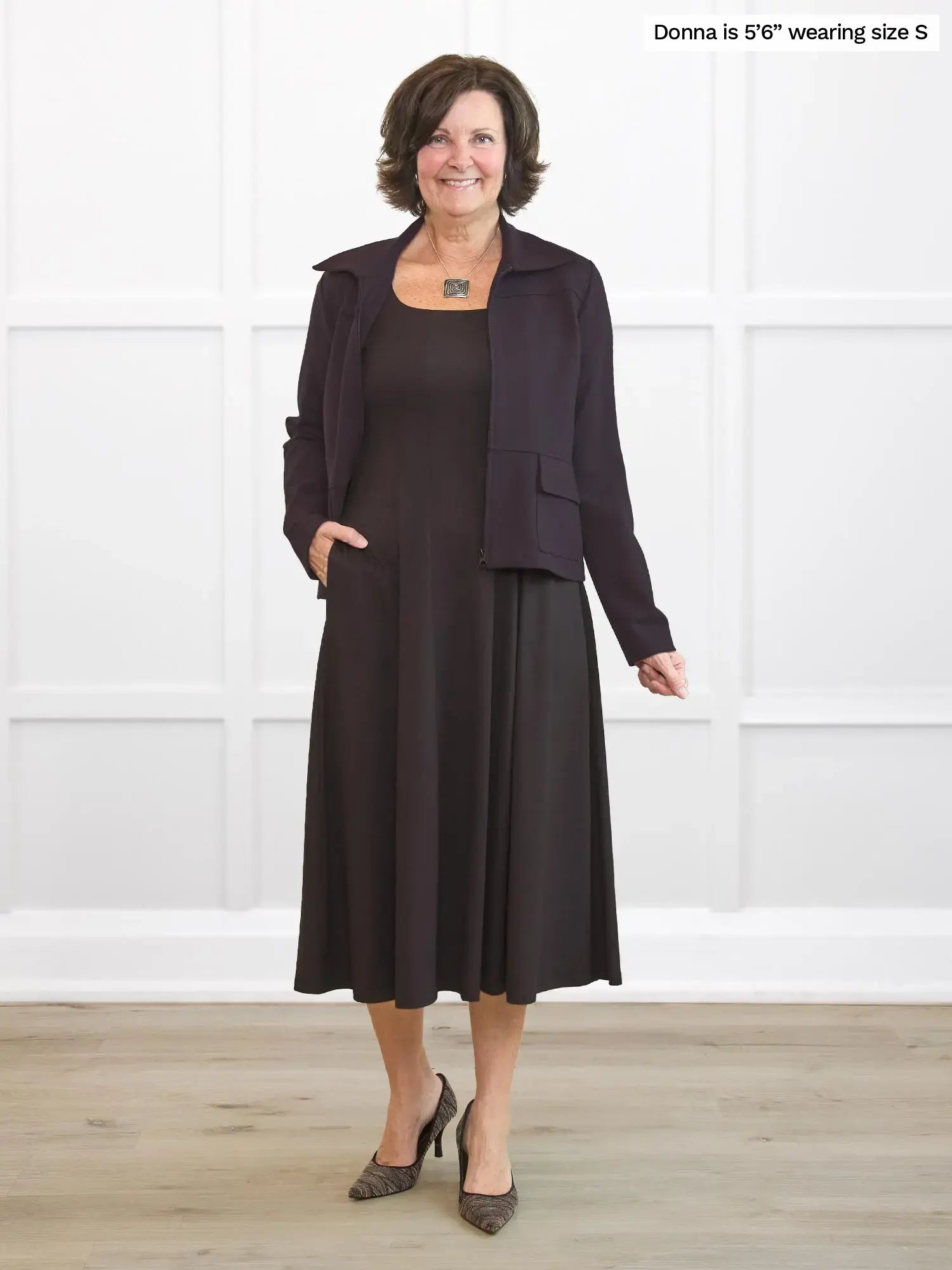 Woman wearing a dark brown dress and jacket set against a white wall.