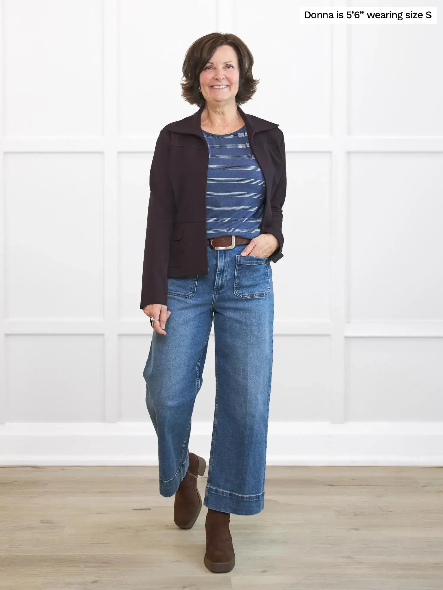 Woman wearing a dark brown jacket, striped shirt, and blue jeans standing against a white paneled wall.