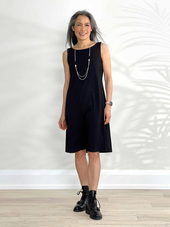 Miik model Lisa (5'6", xsmall) smiling and standing in front of a white wall wearing Miik's Jaaron reversible tank dress in black