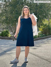 Miik model Christal (5'3", large) smiling wearing Miik's Jaaron reversible tank dress in navy with a white denim jacket over her shoulders and a white converse 