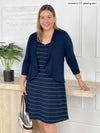 Miik model Christal (5'3", large) smiling while standing next to a table wearing Miik's Jaaron reversible knee length tank dress in navy wide pinstripe with a cropped waterfall cardigan in navy