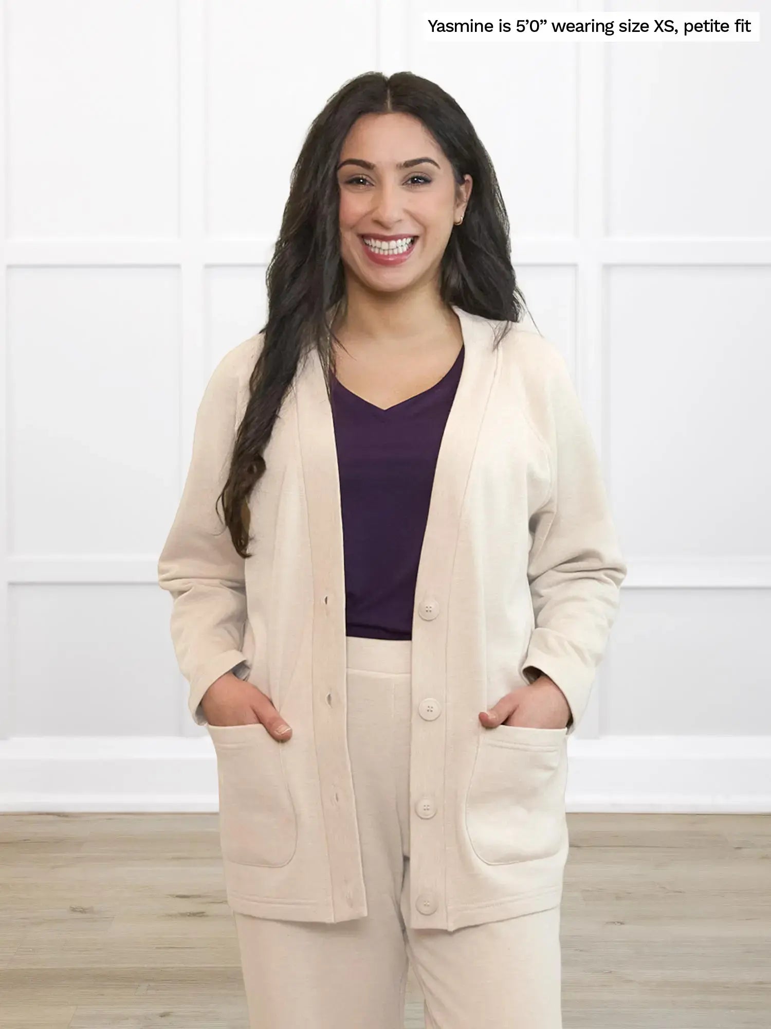 Woman wearing a beige cardigan over a purple top and beige pants in a white room.