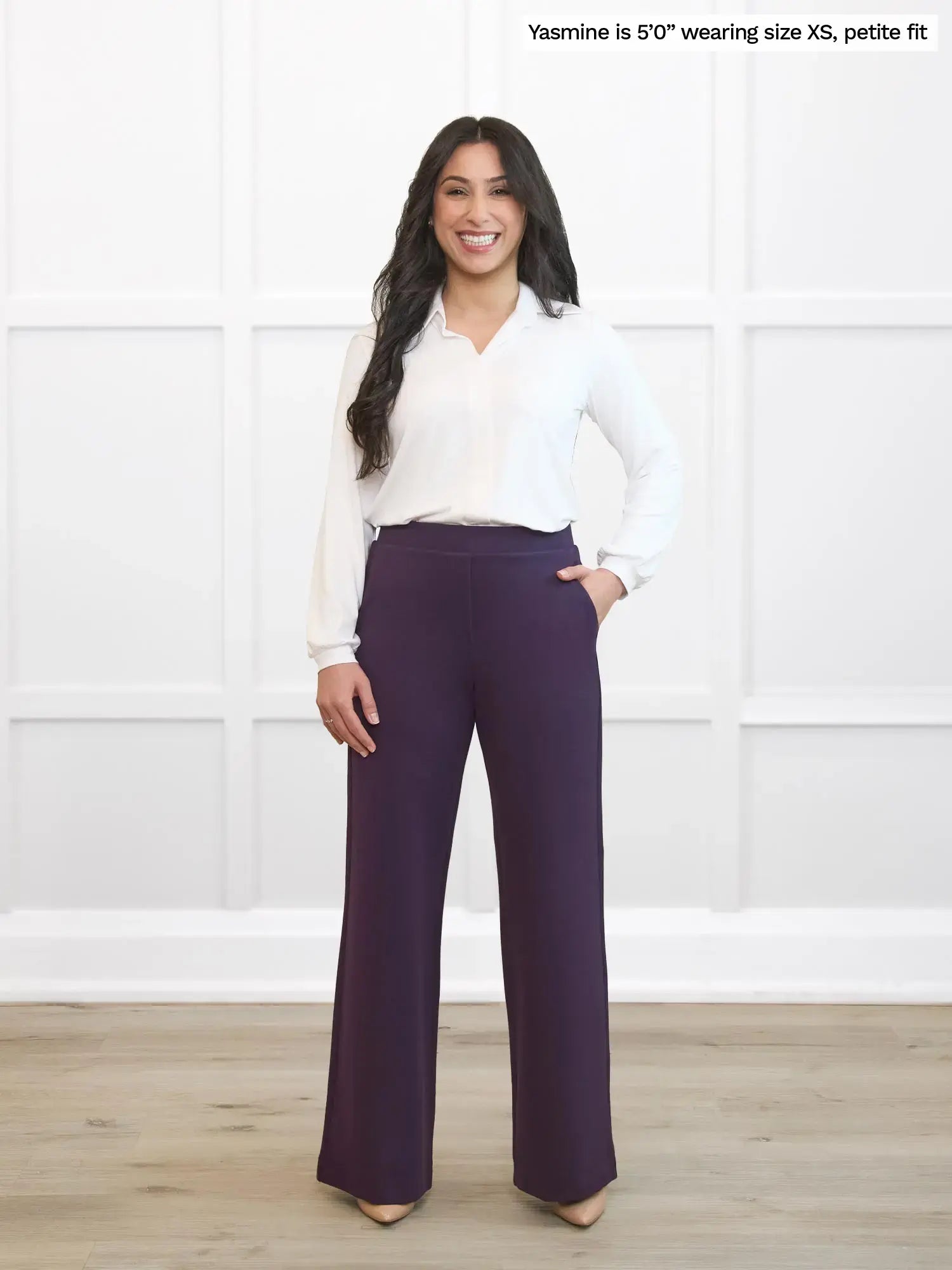 Woman wearing a white blouse and purple pants standing against a white wall.