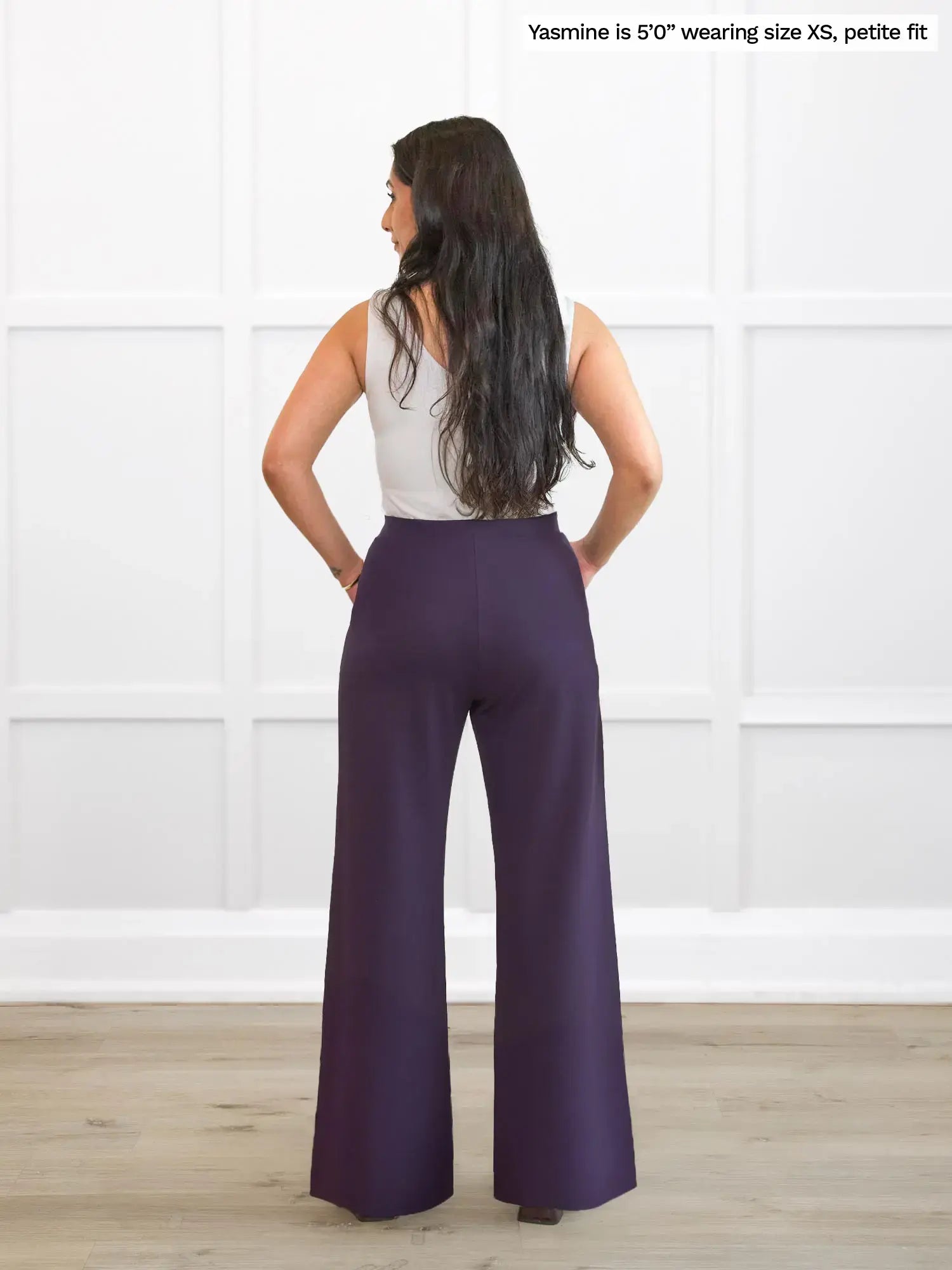 Woman wearing a white top and purple pants against a white wall.