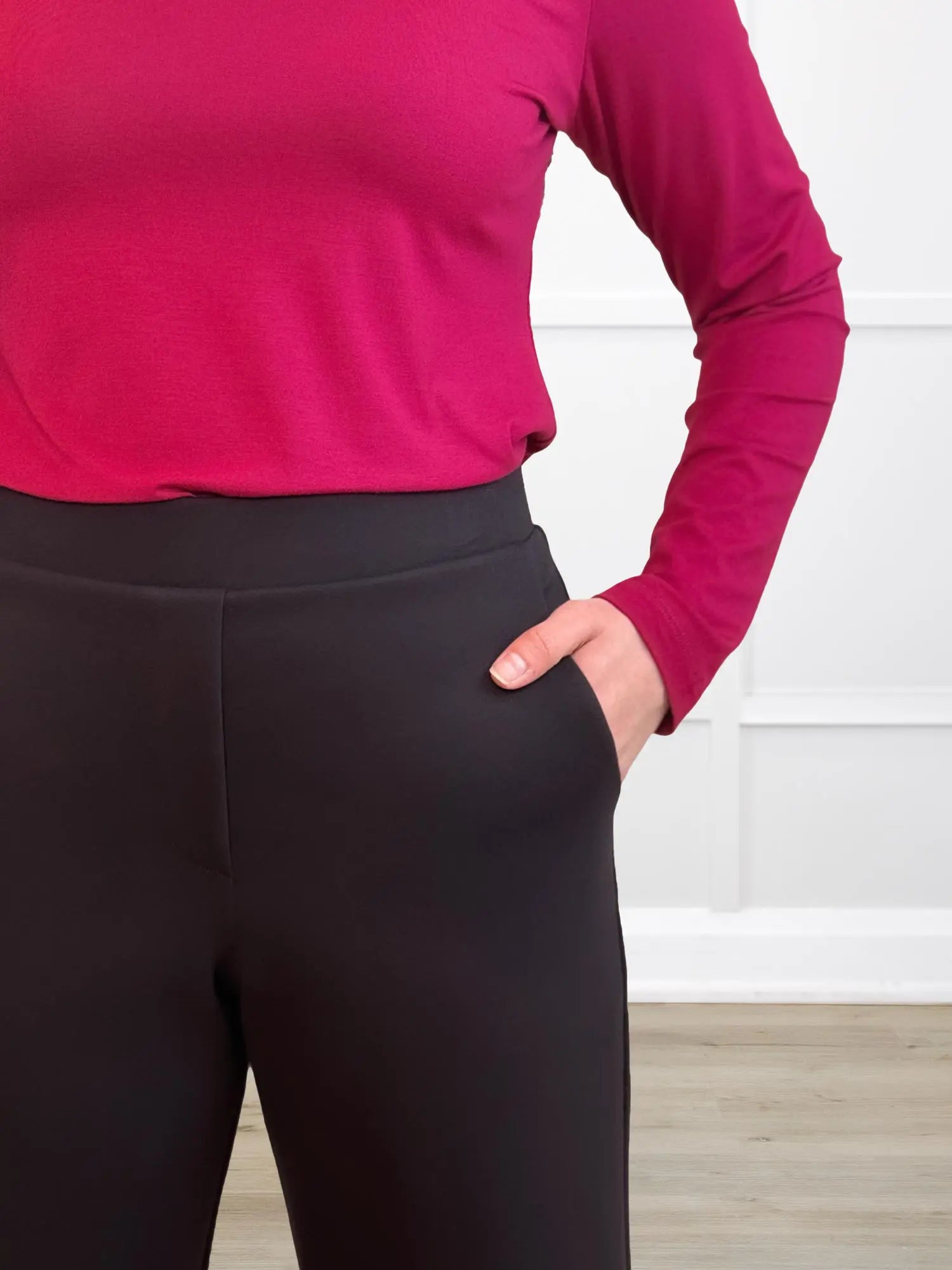 Person wearing a red long-sleeve top and dark brown petite pants against a white wall.