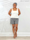 Miik model Keethai (5'5", medium) smiling while leaning against to a white wall wearing Miik's Kavya pull-on casual print shorts in pebble with a square neck tank top in natural