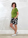 Miik founder Donna (5'6", small) standing sideway in front of a white wall wearing a green moss top with Miik's Kavya pull-on casual print shorts in white lily print 