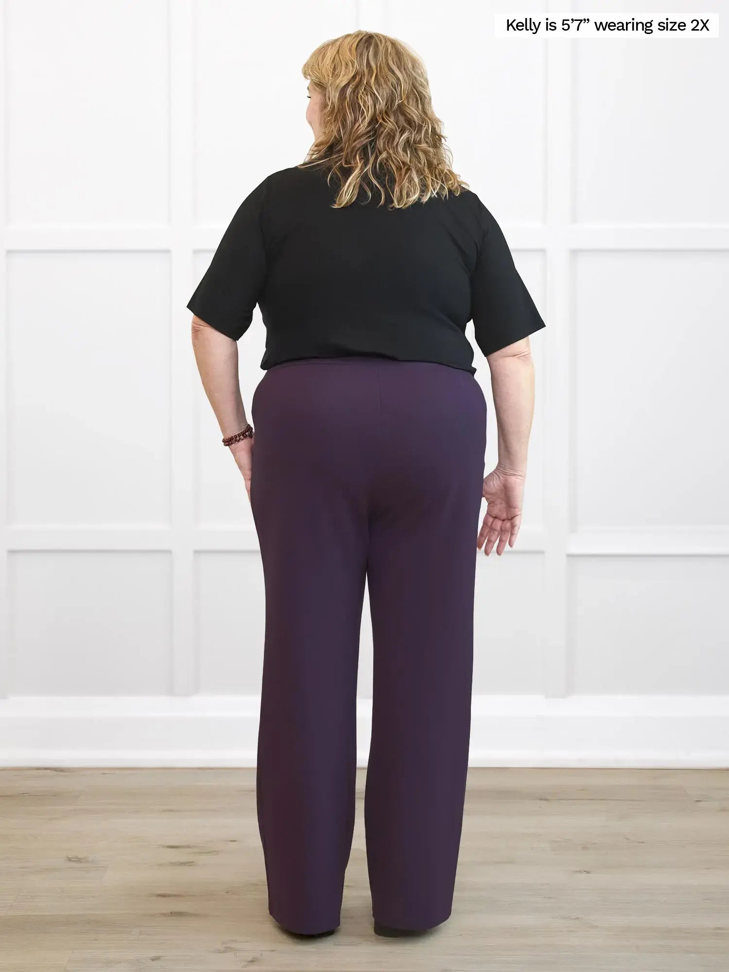 Woman wearing a black top and purple pants against a white wall.