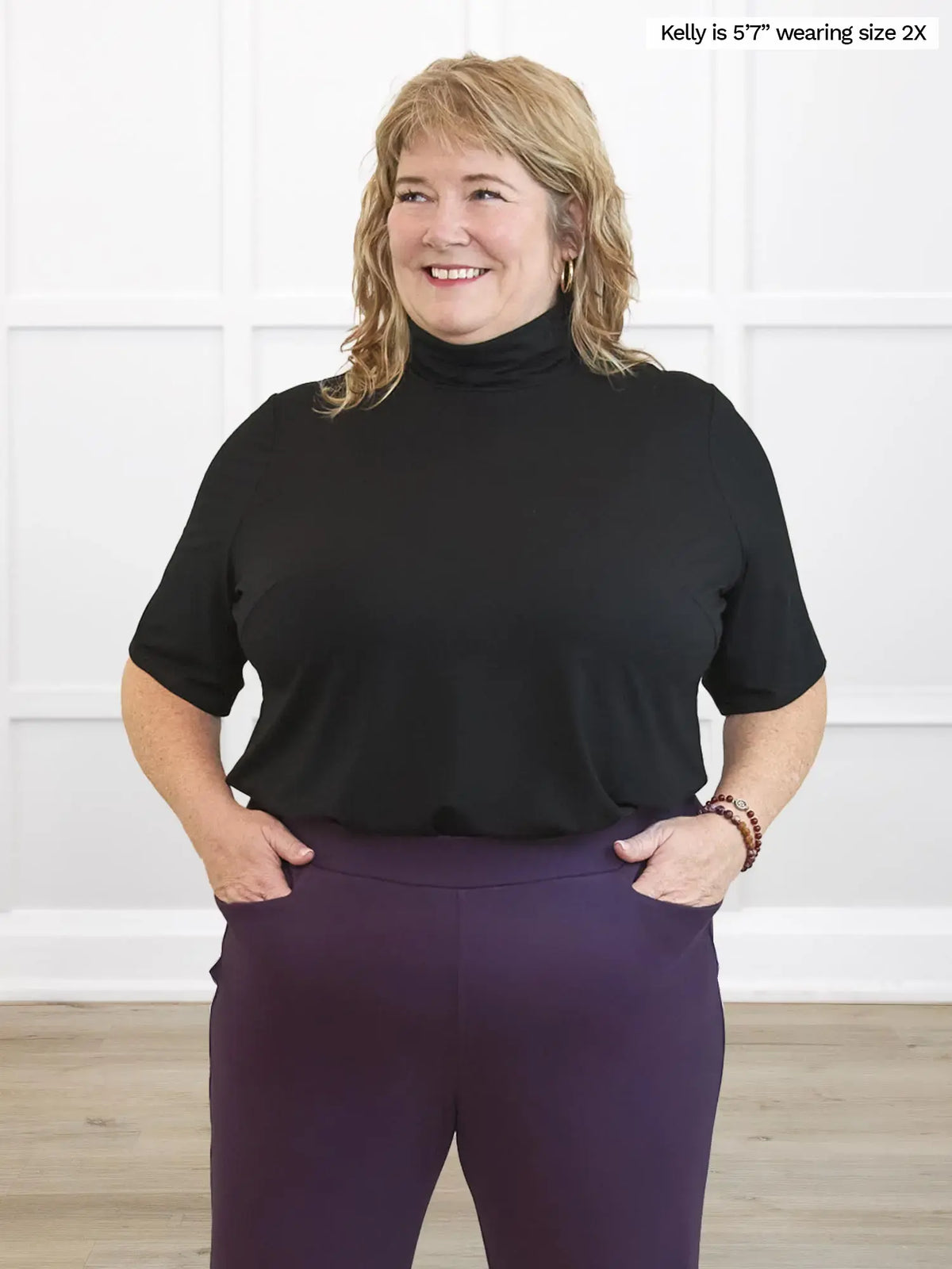 Woman wearing a black turtleneck and purple pants against a white wall.