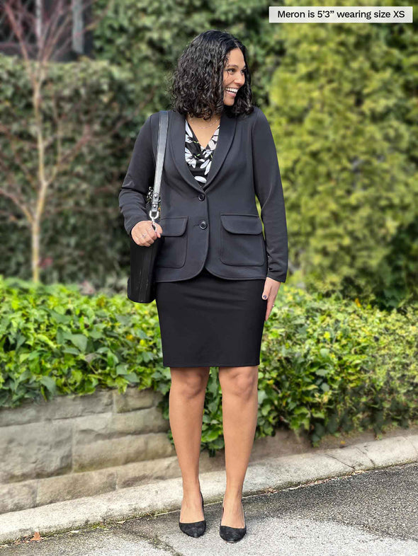 Miik model Meron (5'3", xsmall) smiling and looking away wearing a black pencil skirt along with Miik's Maeve LightLuxe washable blazer in graphite closed 