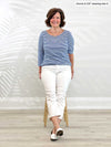 Woman sitting on a stool wearing Miik's Mahala boatneck breton top in blue stripe with white pants.