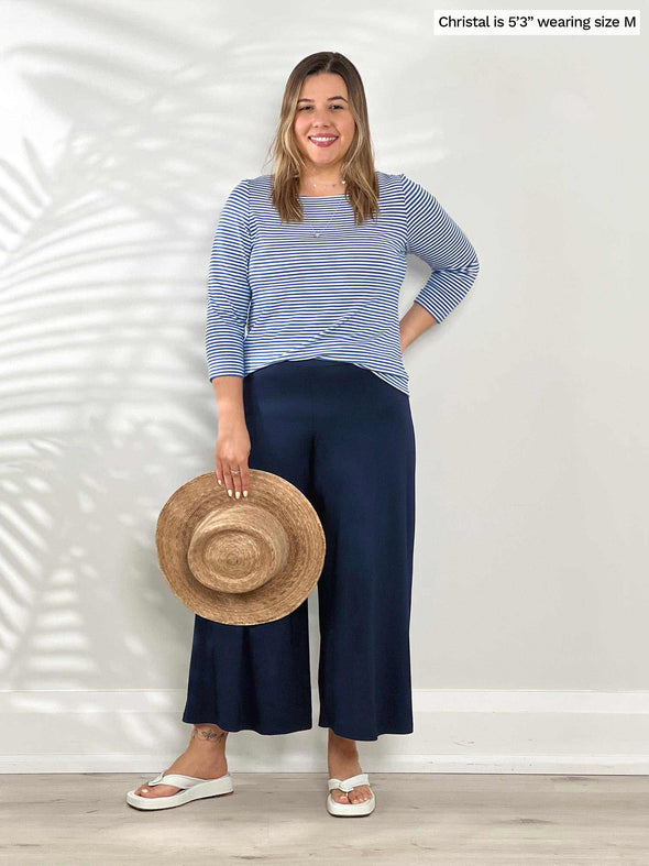 Woman standing in front of a wall wearing Miik's Mahala boatneck breton top in blue stripe with navy capris.