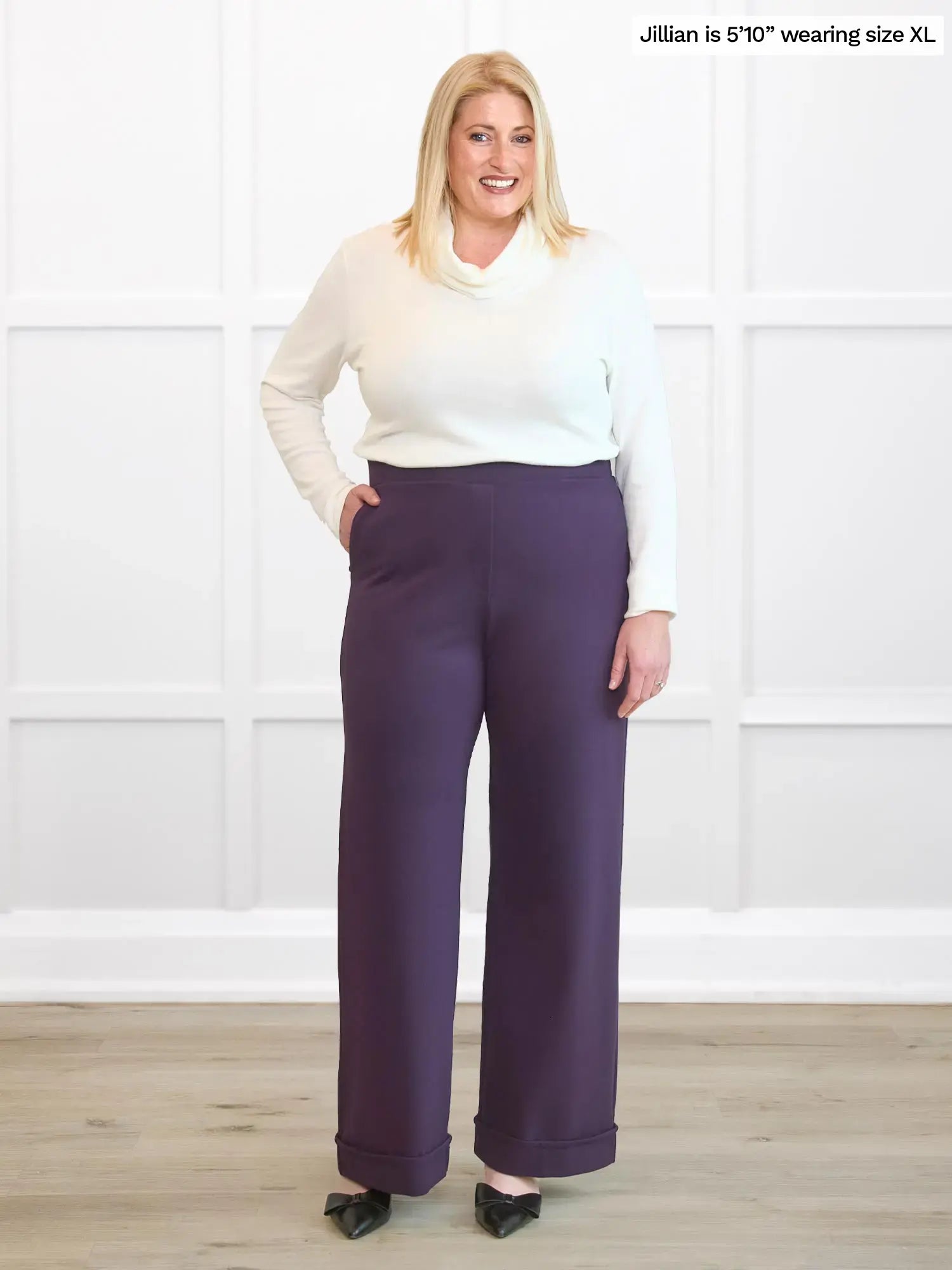 Woman wearing a white top and purple pants against a white wall.