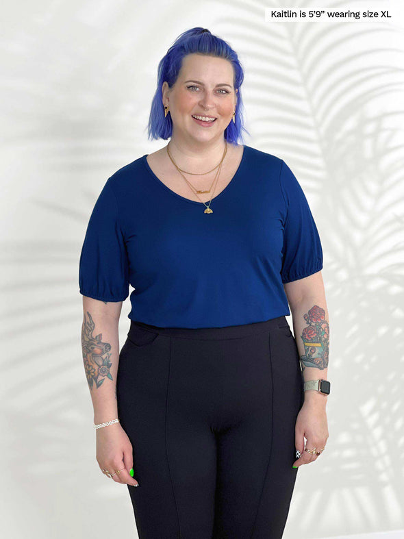 A close up shot of Miik model Kaitlin (5'9", xlarge) smiling wearing Miik's Makena v-neck puff sleeve blouse in ink blue tucked in a black dress pant 