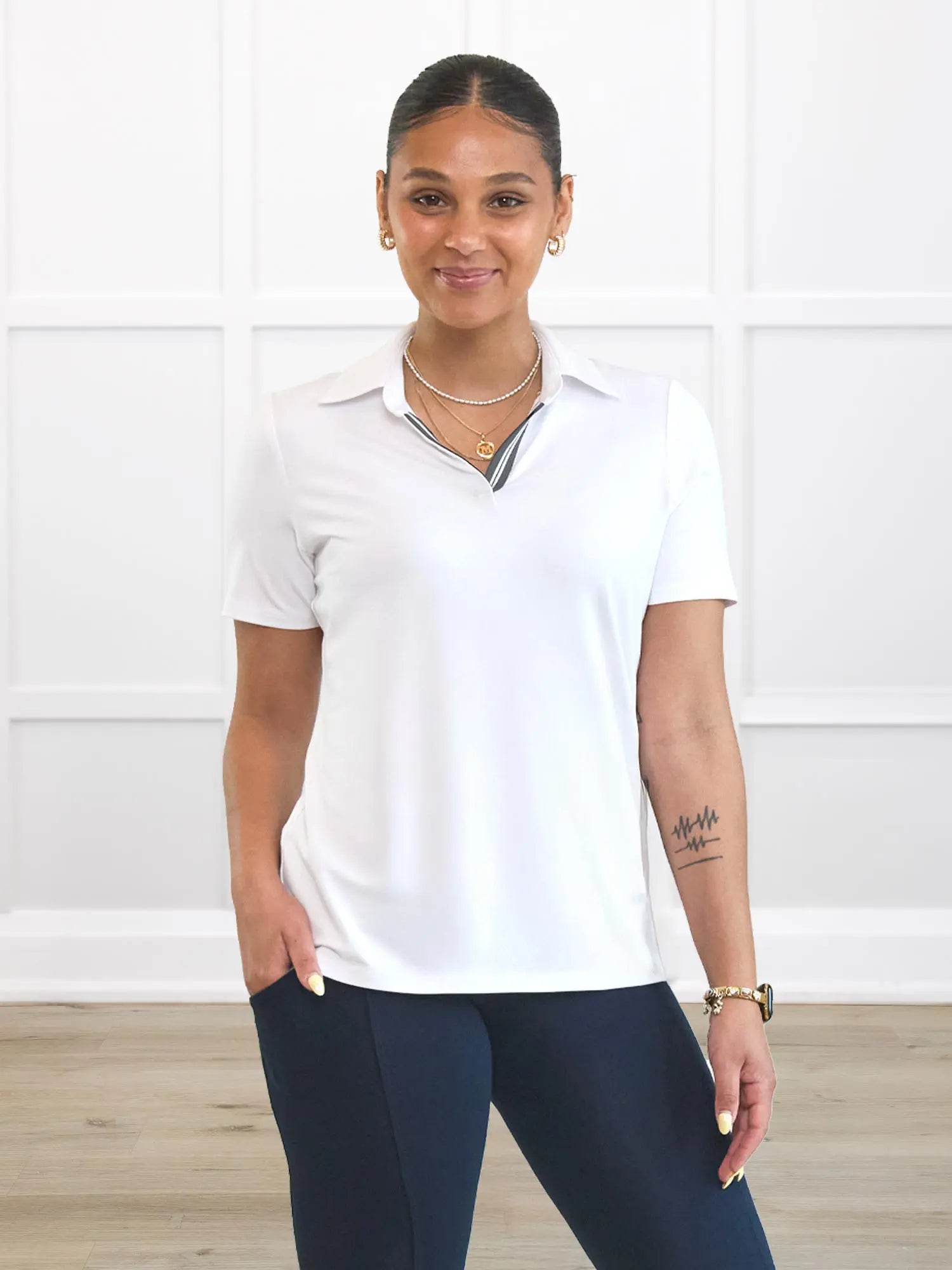 A woman modeling a white short-sleeve polo shirt with a dark gray and white striped inner placket, paired with navy blue leggings, standing in front of a white paneled wall.