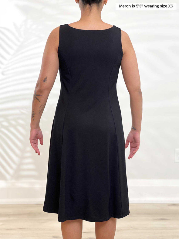 Miik model Meron (5’3”, xsmall) standing with her back towards the camera showing the back of Miik's Niah reversible knee length flounce dress in black