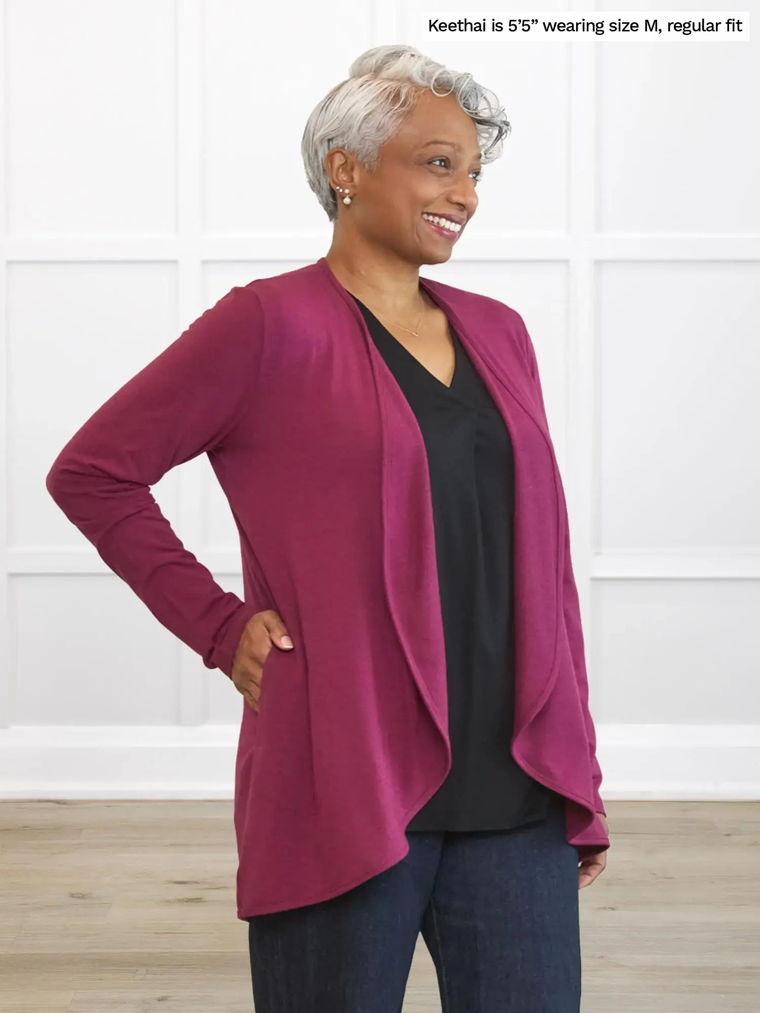 Woman wearing a magenta cardigan over a black top and blue jeans, standing against a white wall.