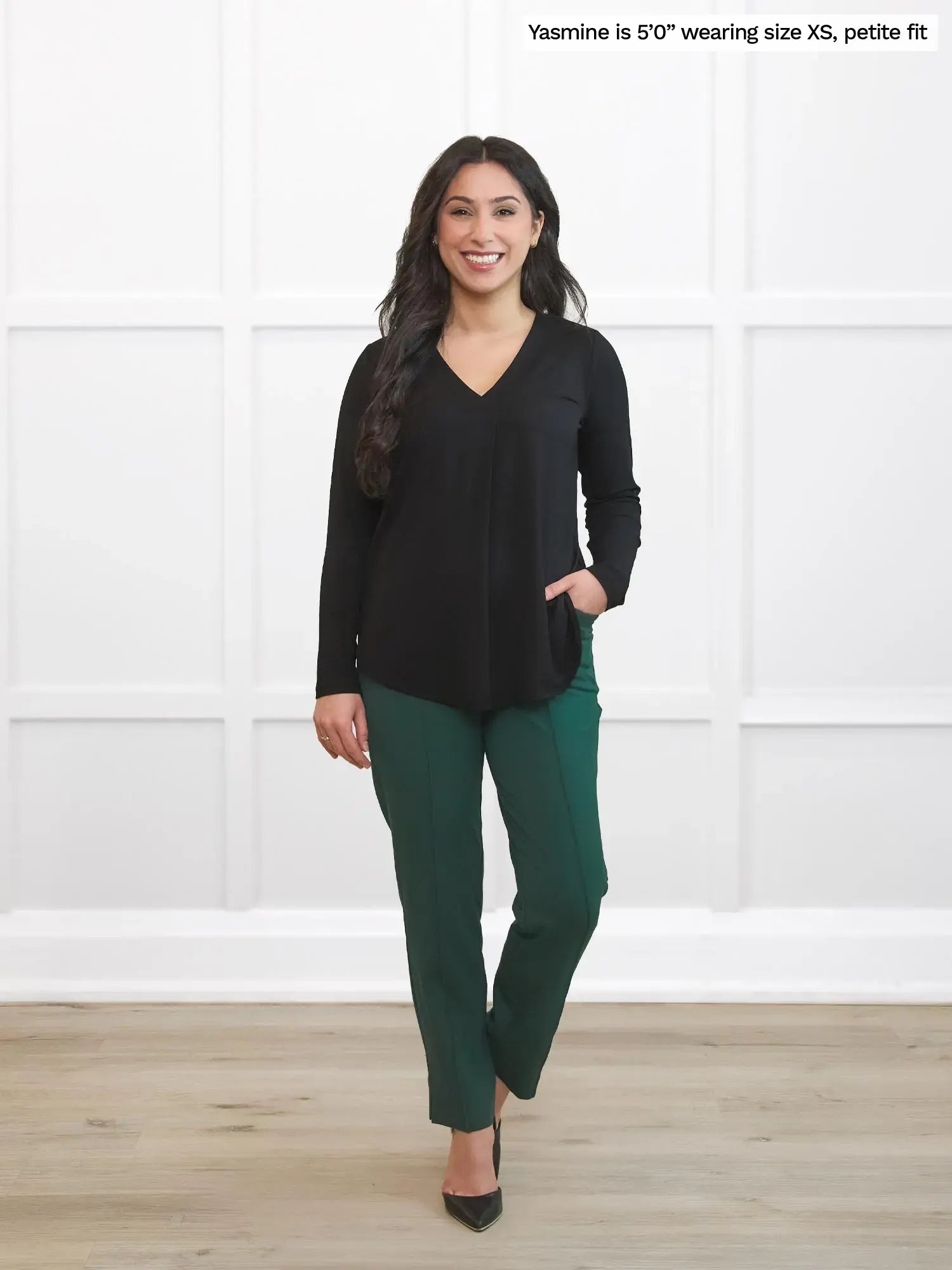 Woman wearing a black top and green pants standing against a white wall.