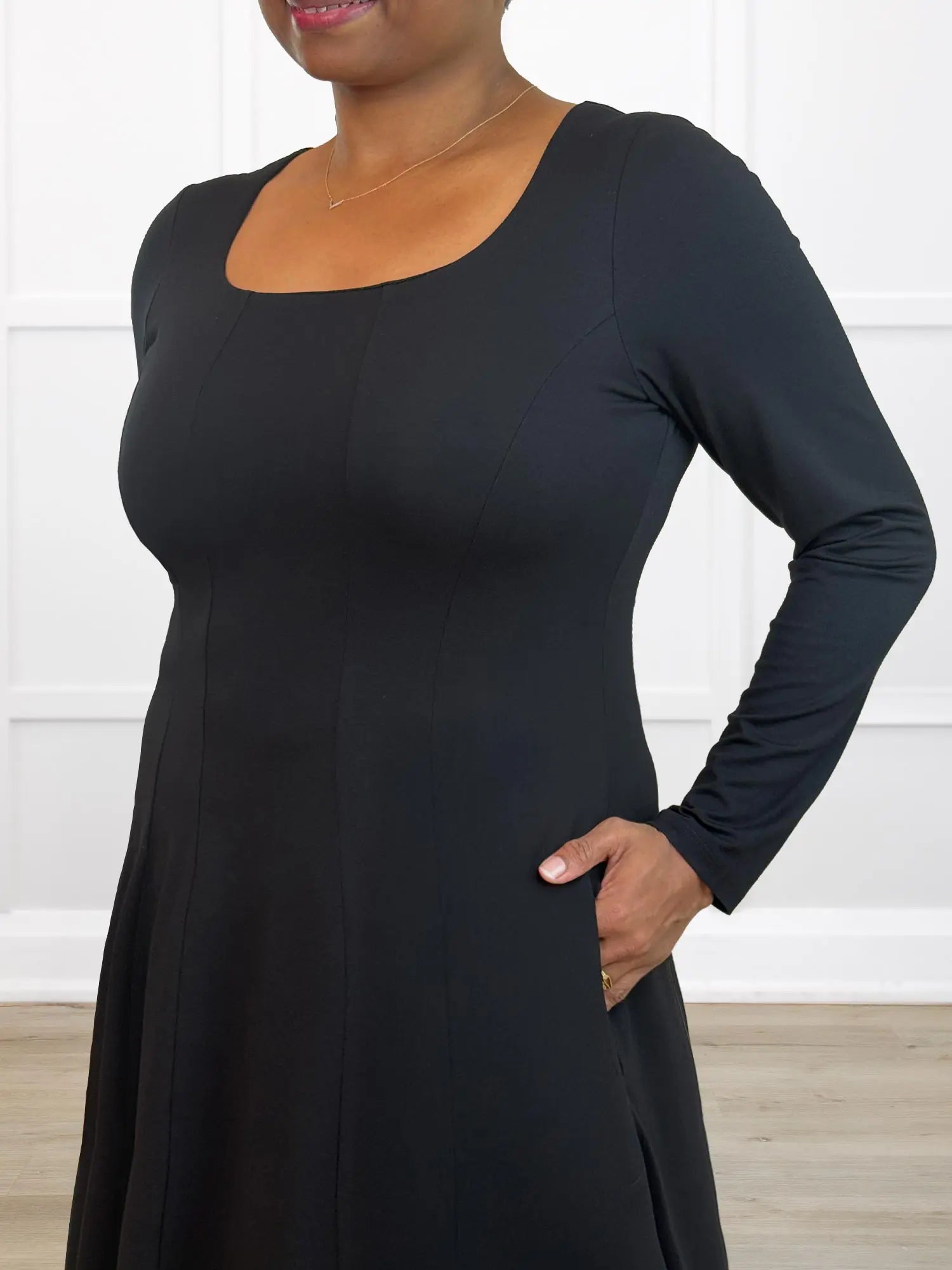Woman wearing a black long-sleeve dress against a white wall.