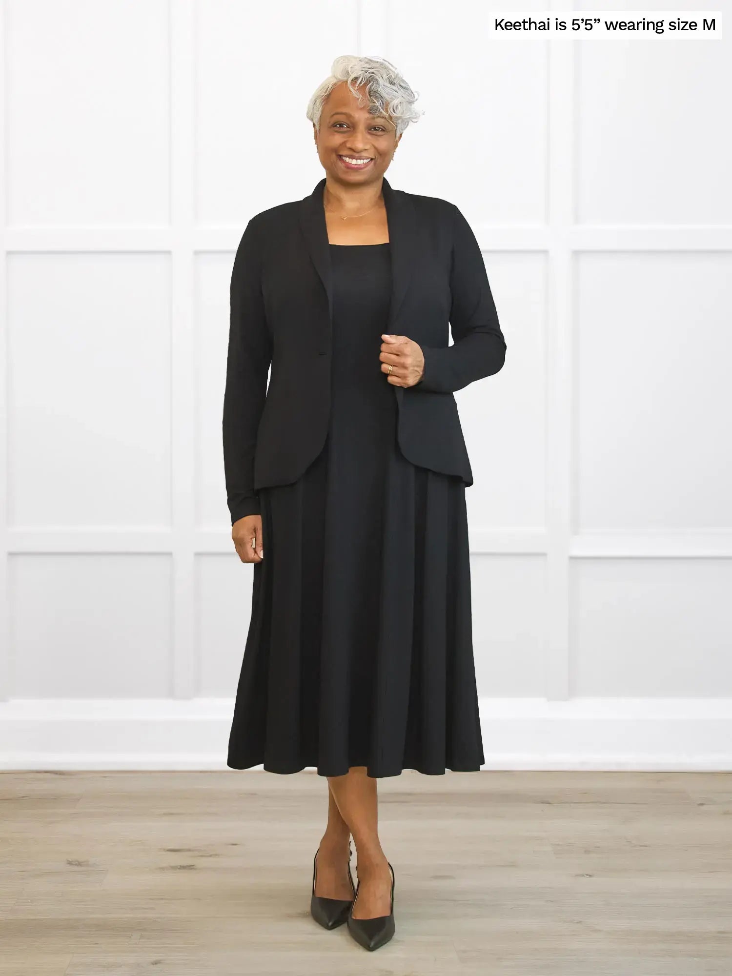Woman wearing a black blazer over a black dress standing against a white wall.