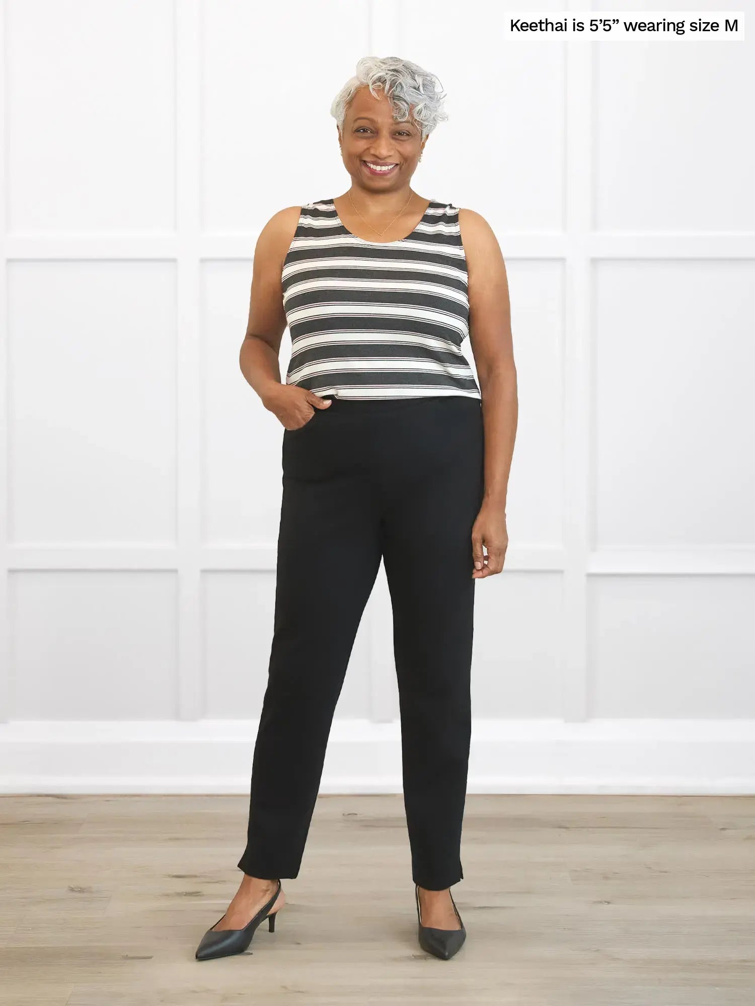 Woman wearing a striped sleeveless top and black pants standing against a white wall.
