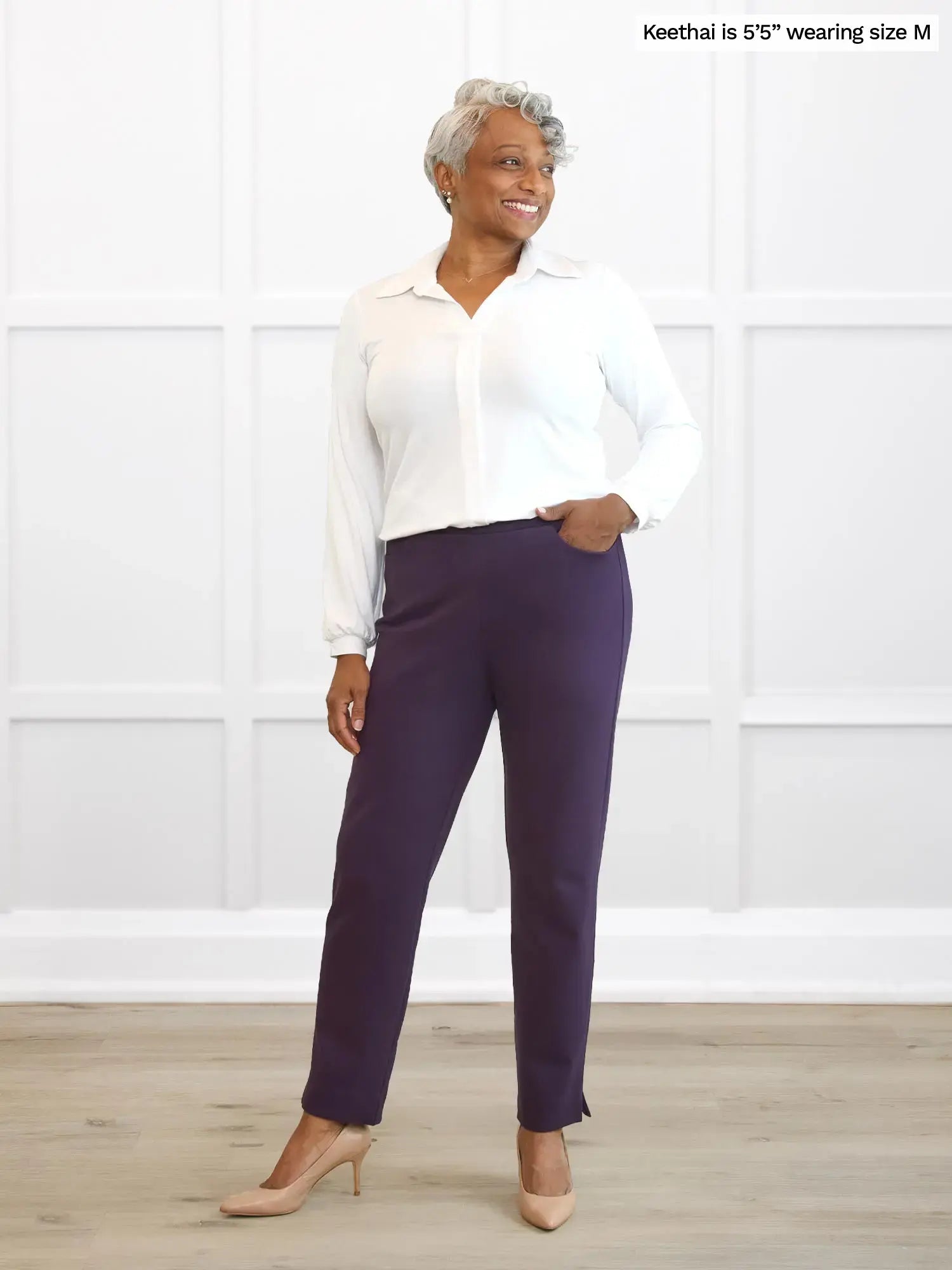 Woman wearing a white blouse and purple pants standing against a white paneled wall.