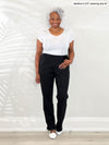 Miik model Keethai (5'5", medium) smiling while standing in front of a white wall wearing a white basic tee along with Miik's Roma pull-on straight leg ankle pant in black