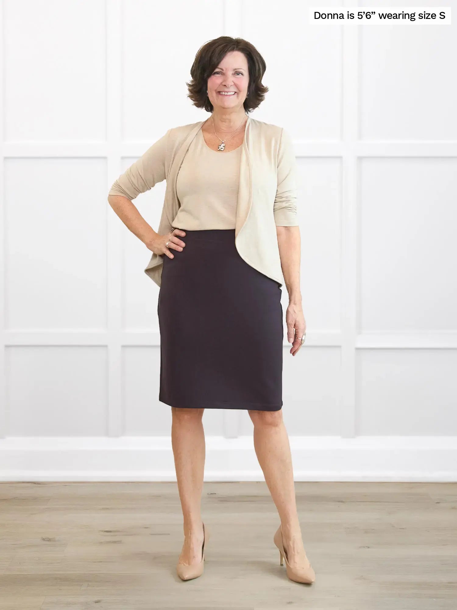 Woman wearing a beige cardigan, dark brown skirt, and beige heels in a white room.