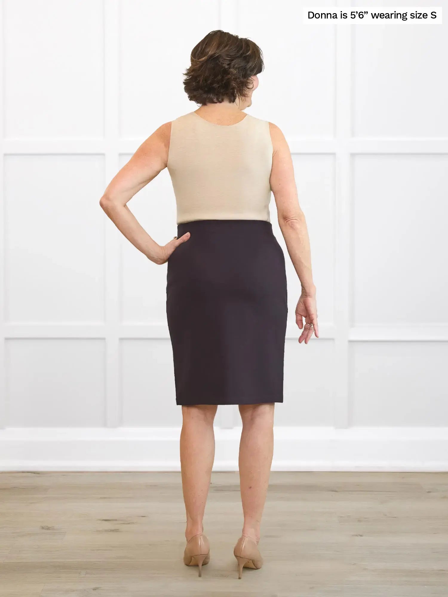 Woman wearing a beige sleeveless top and dark brown skirt standing against a white wall.
