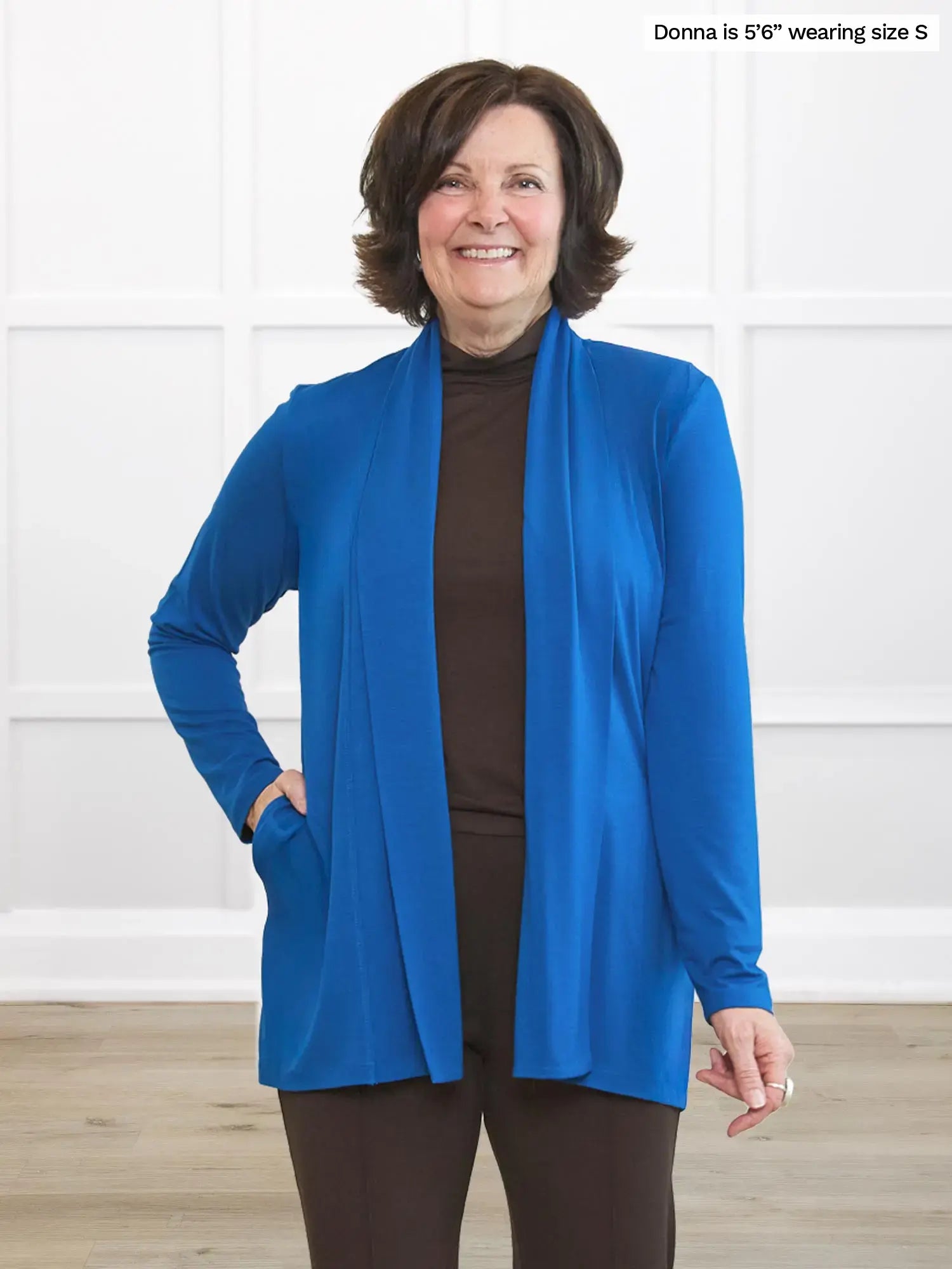 Woman wearing a blue cardigan over a brown top and pants, standing against a white wall.