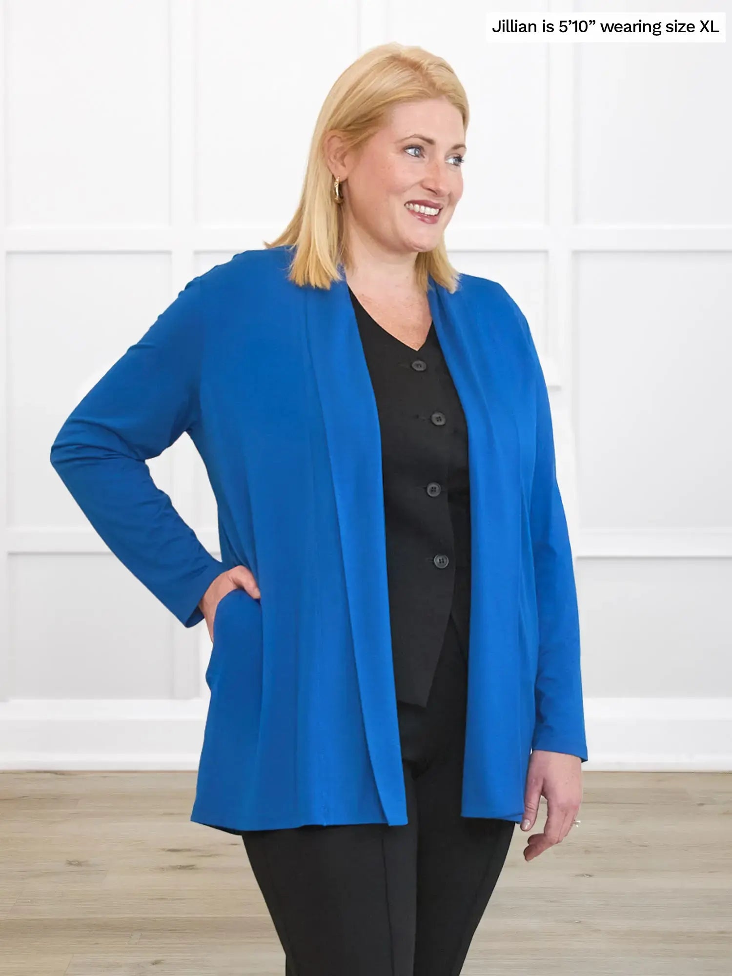 Woman wearing a blue cardigan over a black top and pants, standing against a white wall.
