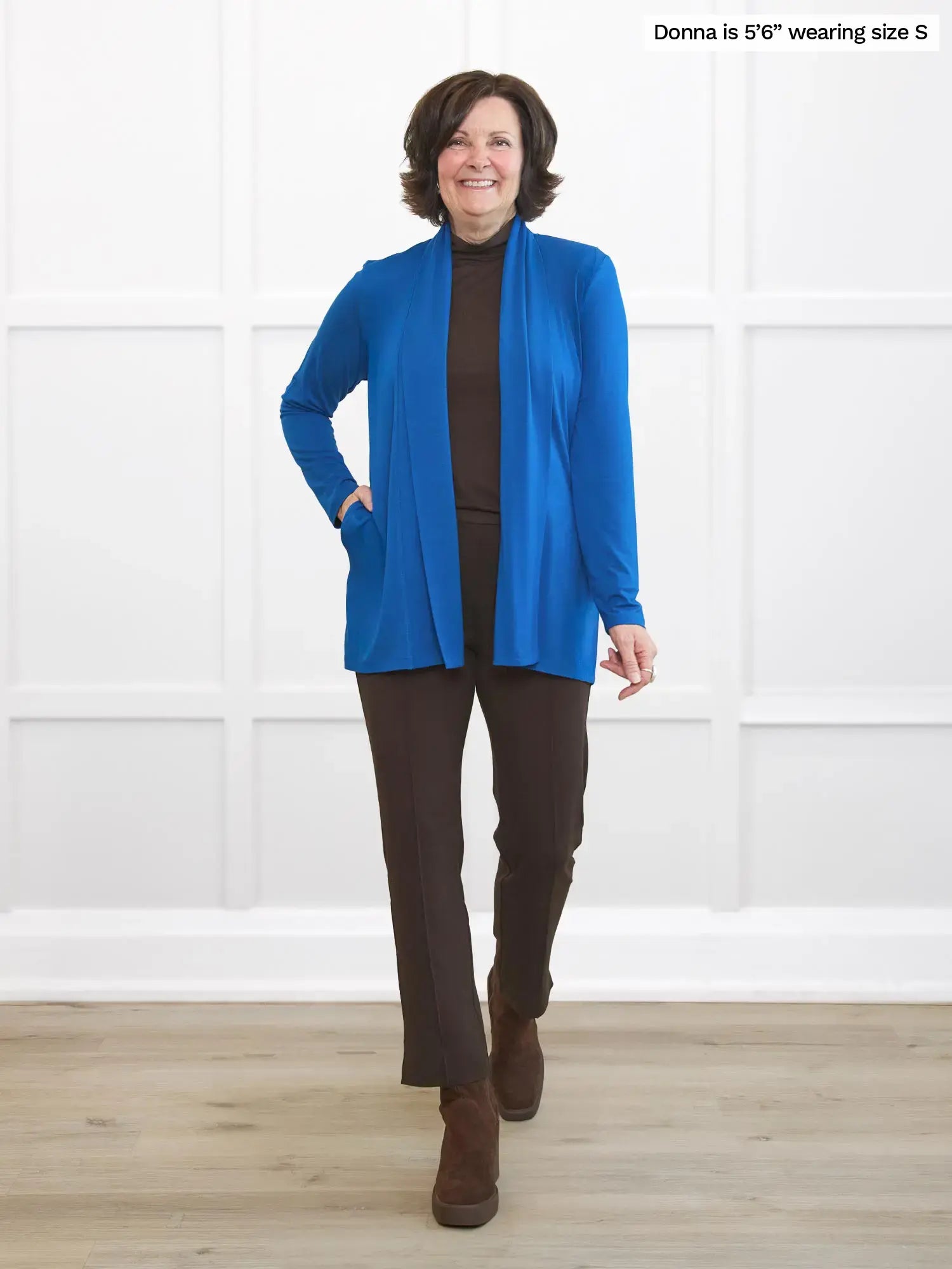 Woman wearing a blue cardigan, brown pants, and brown boots standing against a white wall.