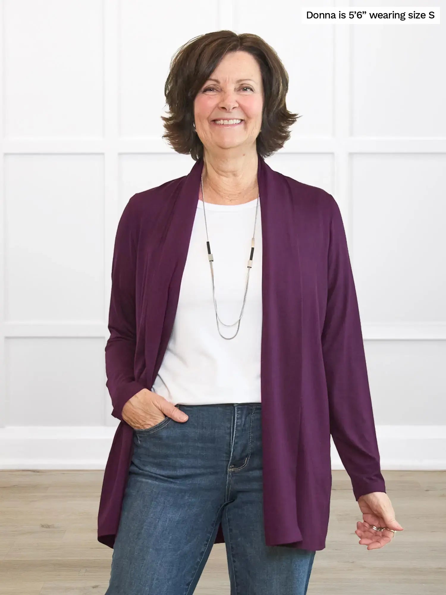 Woman wearing a purple cardigan over a white shirt and blue jeans, standing against a white background.