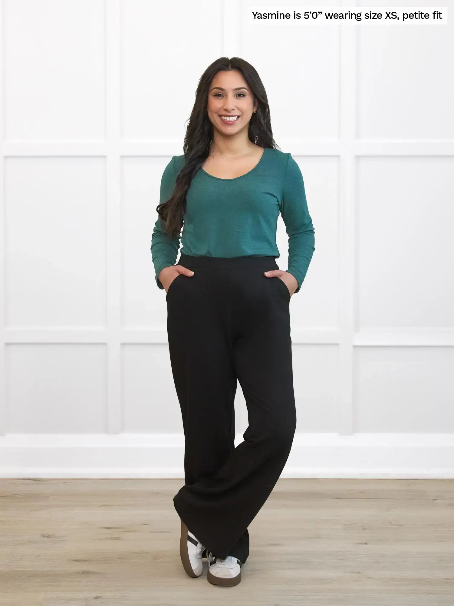 Woman wearing a teal top and black pants standing against a white wall.