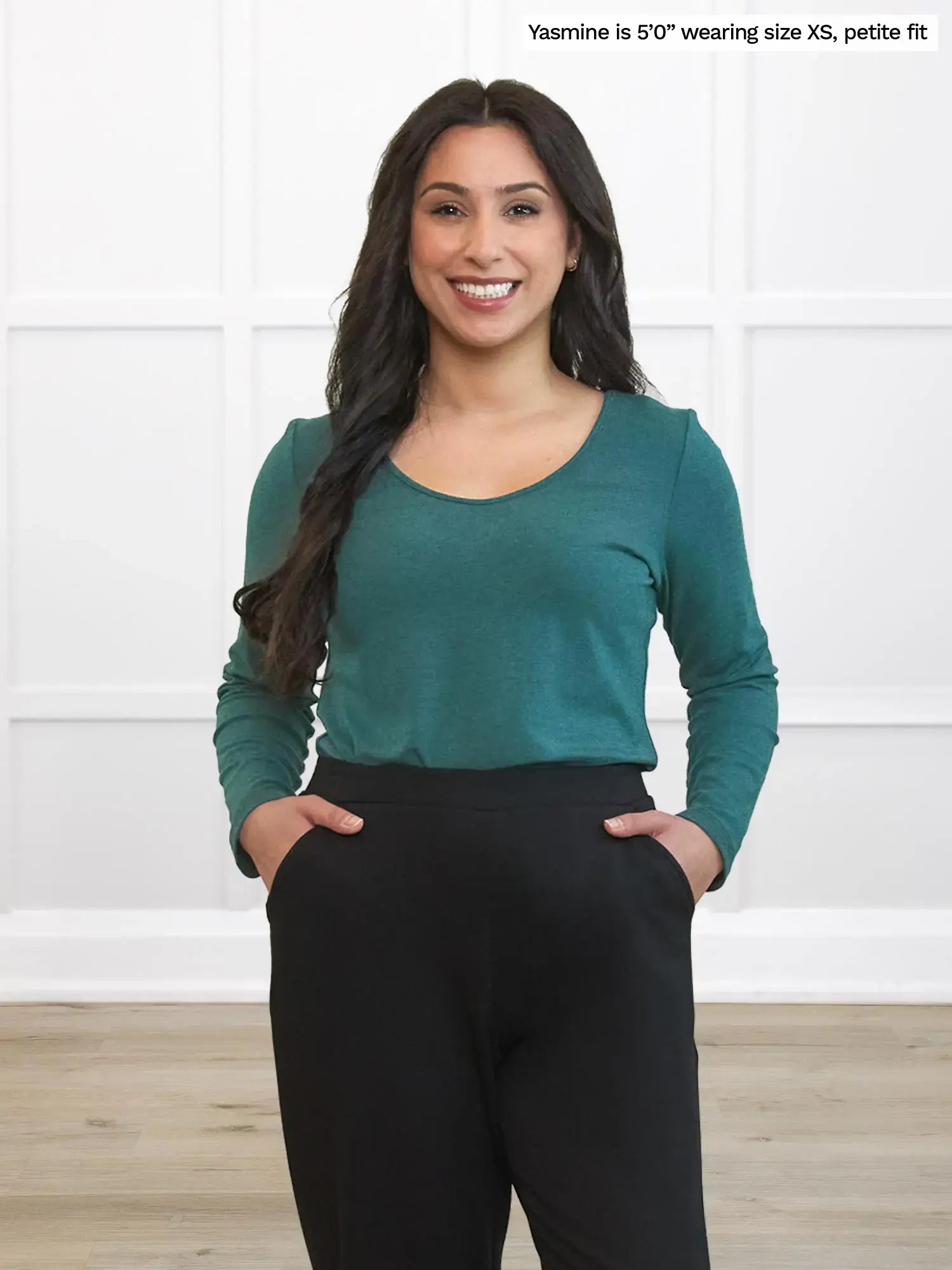 Woman wearing a green long-sleeve top and black pants against a white wall.
