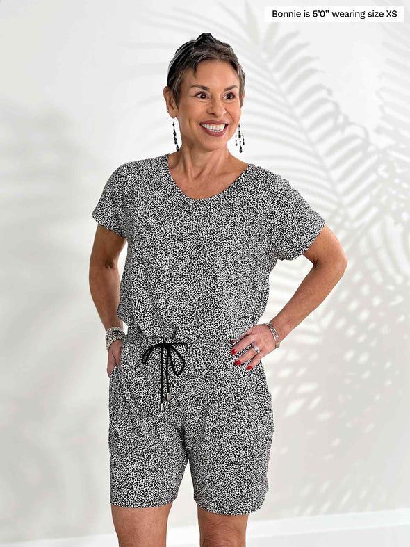 Miik model Bonnie (5'0", xsmall, petite) smiling while standing in front of a white wall wearing Miik's Tanya short sleeve open-back romper in pebble print 